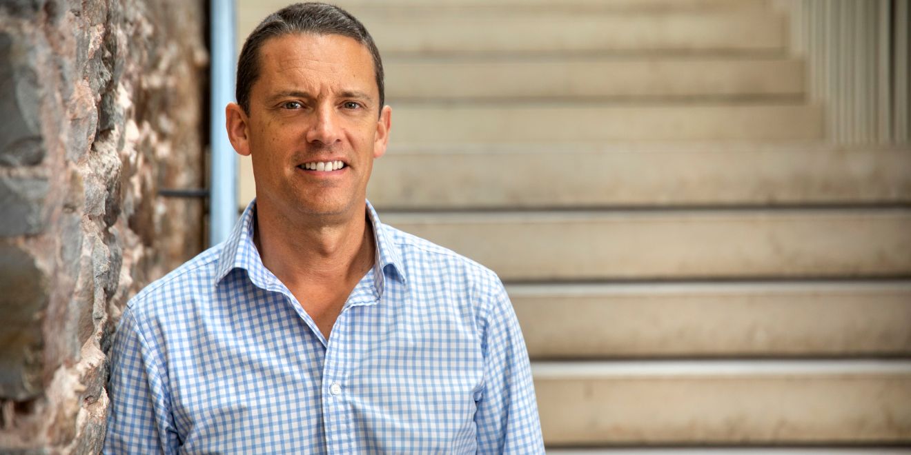 A headshot of a man with short brown hair wearing a blue and white checked shirt facing the camera