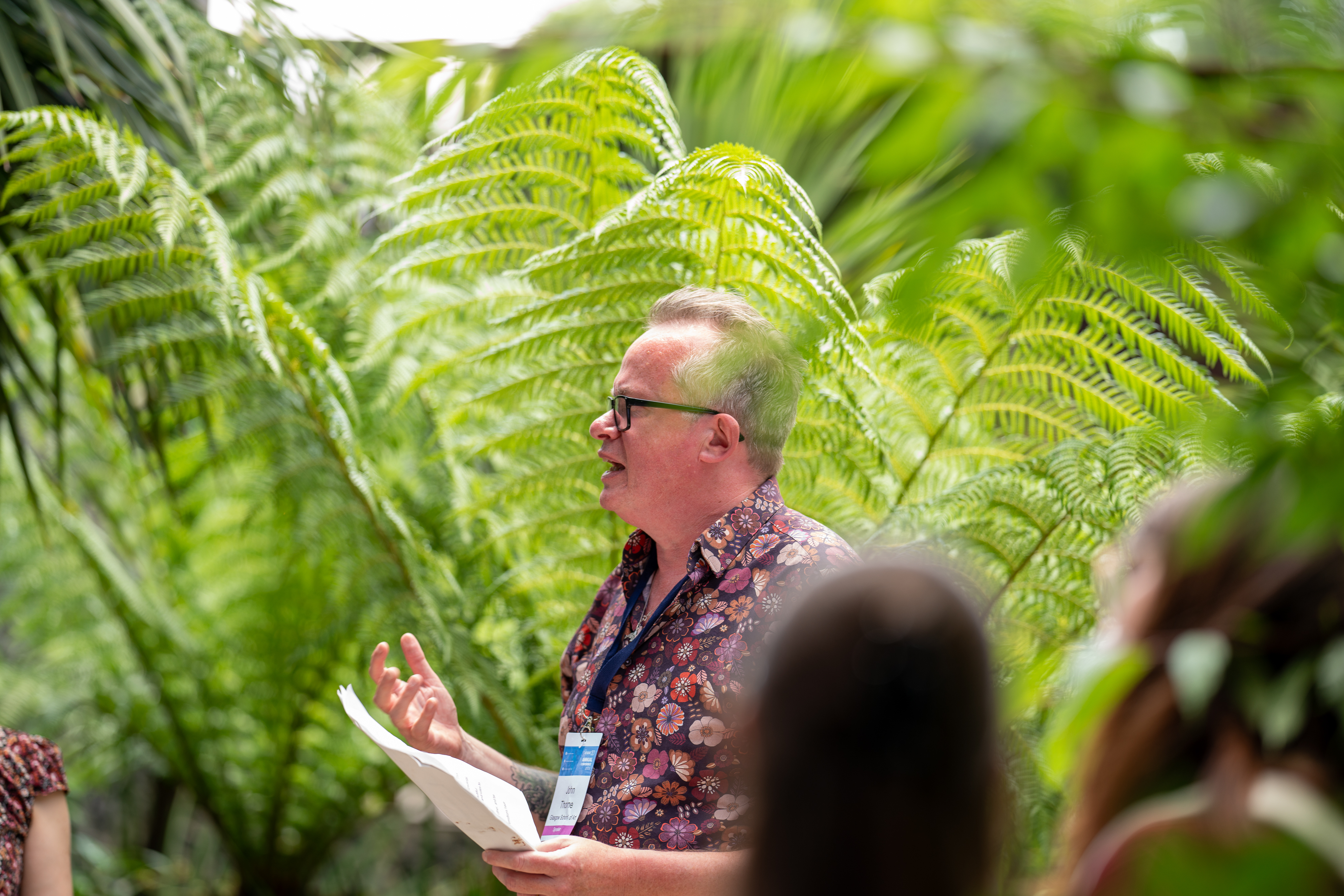 A man wearing a lanyard and a flowery shirt stands in front of a large lush green fern trees.