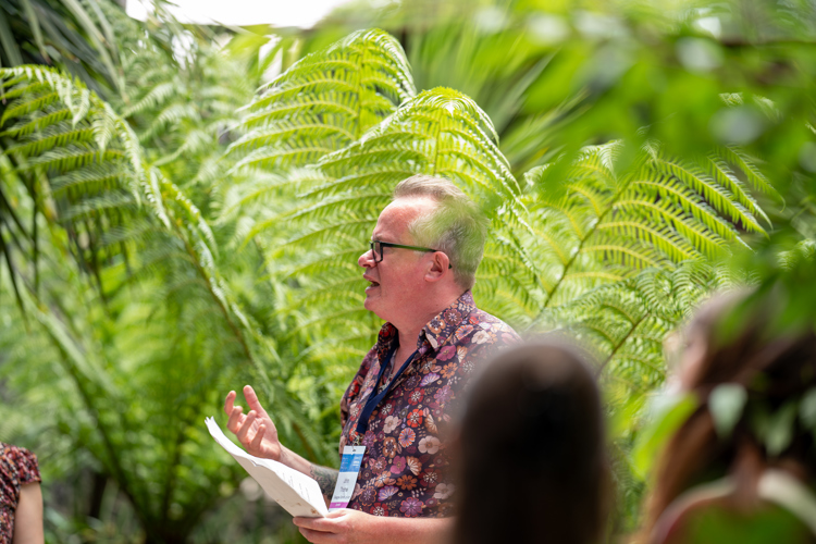  A man stands wearing a patterned shirt stands in front of lush green palm trees 